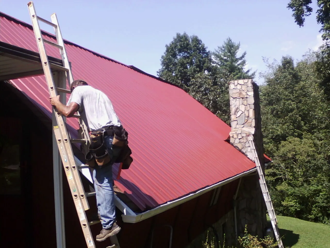 Roofer on a ladder beside a red metal roof and stone chimney — Soffit & Fascia Repair by Roofing Brothers in Elizabethton